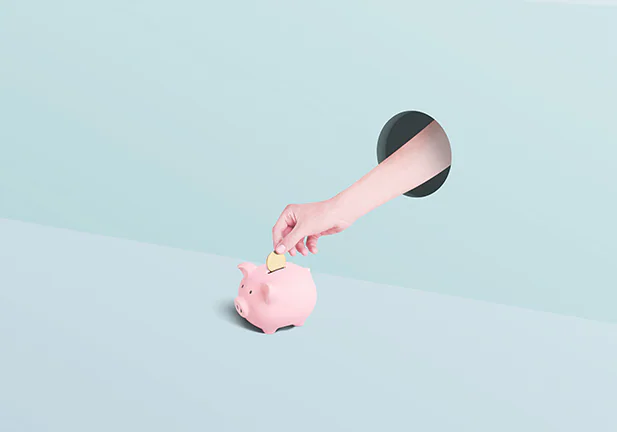 Human hand sticking through a hole in the wall placing a gold coin into a pink piggy bank on a greenish two toned background/