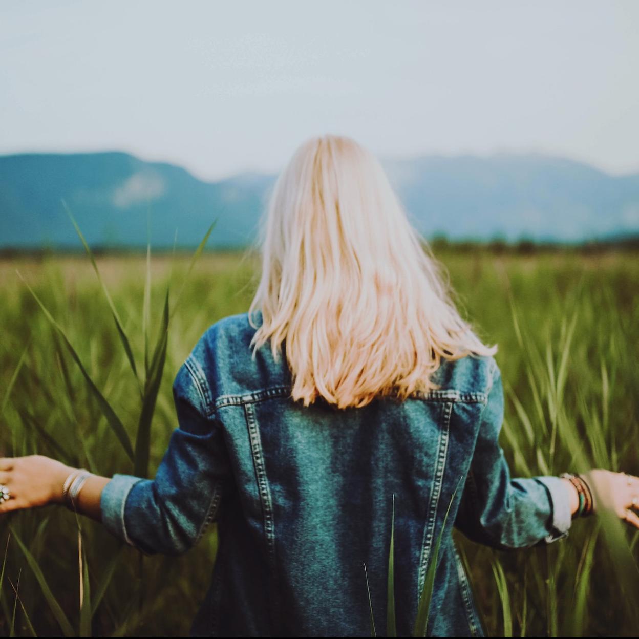 Mujer rubia con cazadora vaquera paseando por un campo verde. /Foto de Tobi, en Pexels.