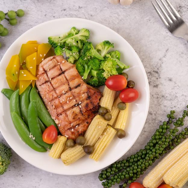 Plato con un filete de pollo cubierto con sésamo blanco, guisantes, tomates, brócoli y calabaza. 