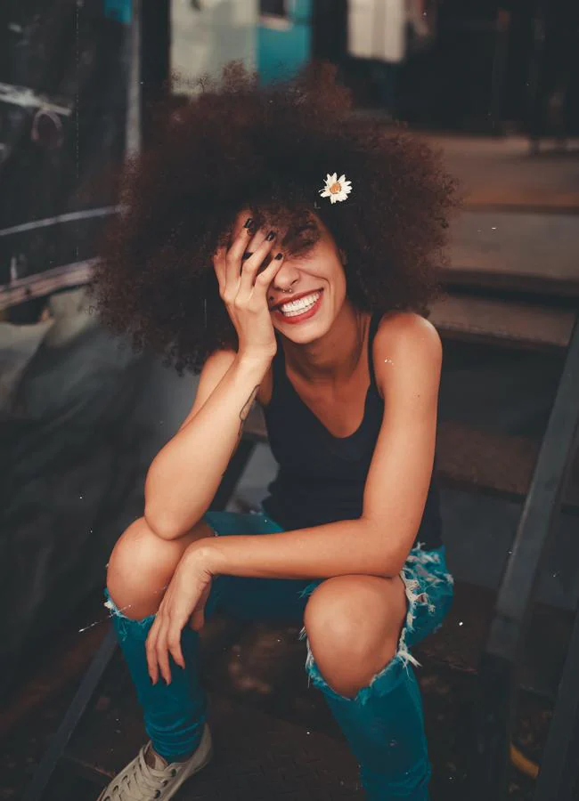 Mujer con el pelo afro sonriendo y sentada en unas escaleras. / Foto de luizclas en Pexels. 